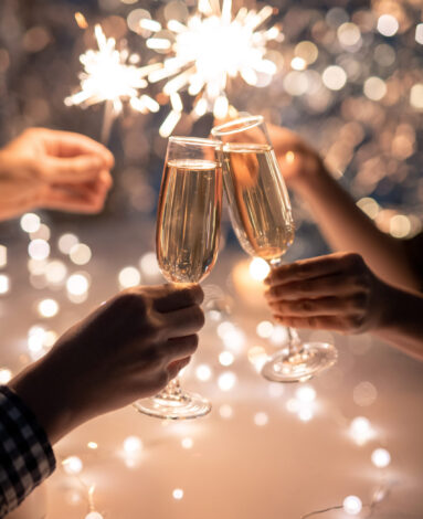 Hands of young couple clinking with flutes of champagne on background of two humans holding sparkling bengal lights