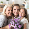 Headshot portrait of happy three generations women family celebrating mothers day birthday together, smiling grandmother and kid daughter congratulating young mom with flowers gift looking at camera